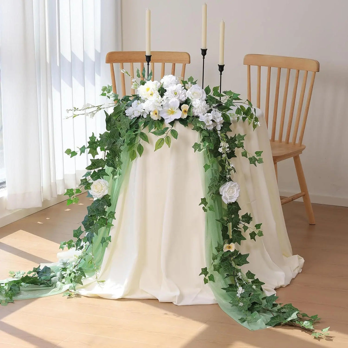 A white and green artificial ivy garland draped over a light-colored fabric, decorated with white flowers, next to wooden cha