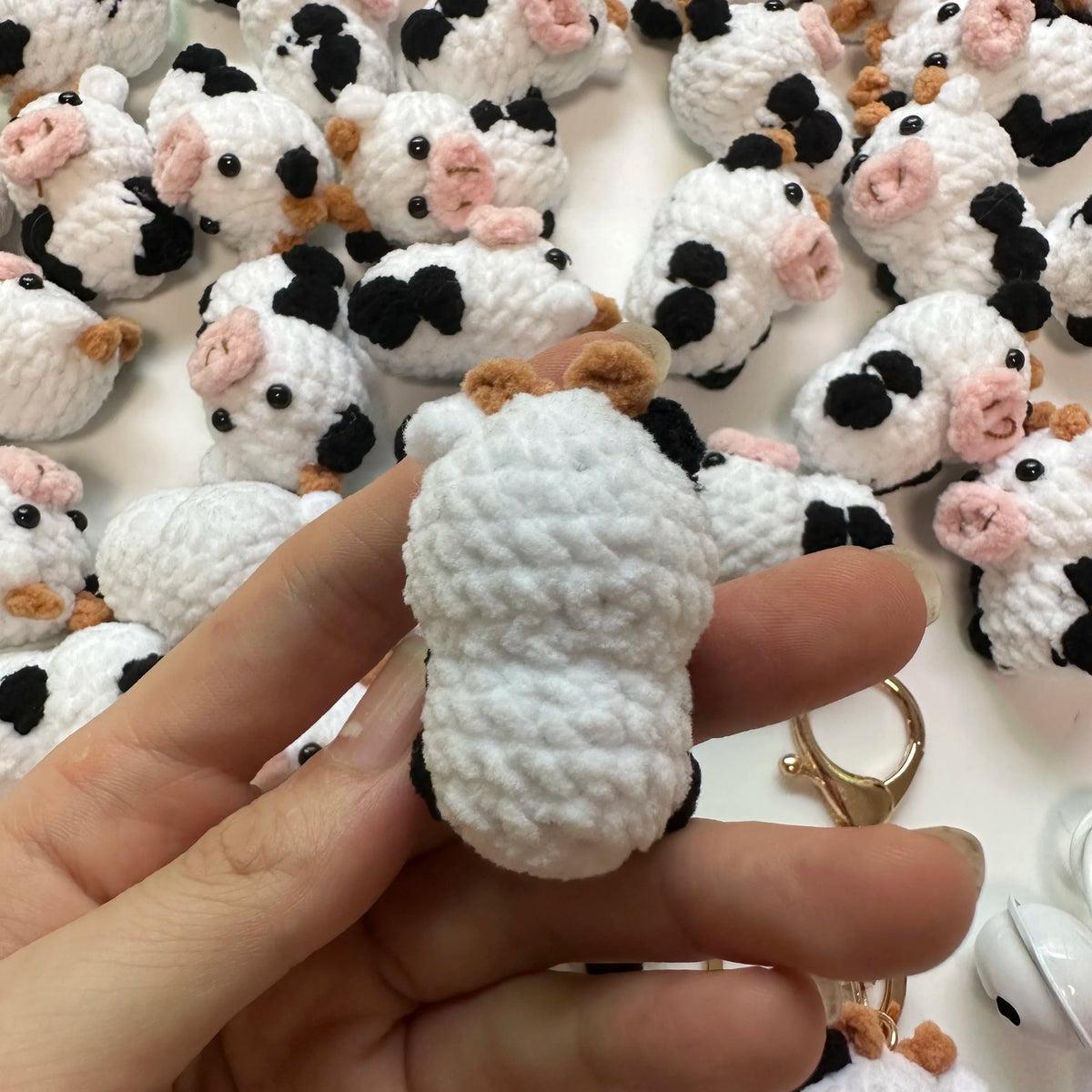 A hand holds a handmade crochet cow keychain featuring white and black wool with pink accents.