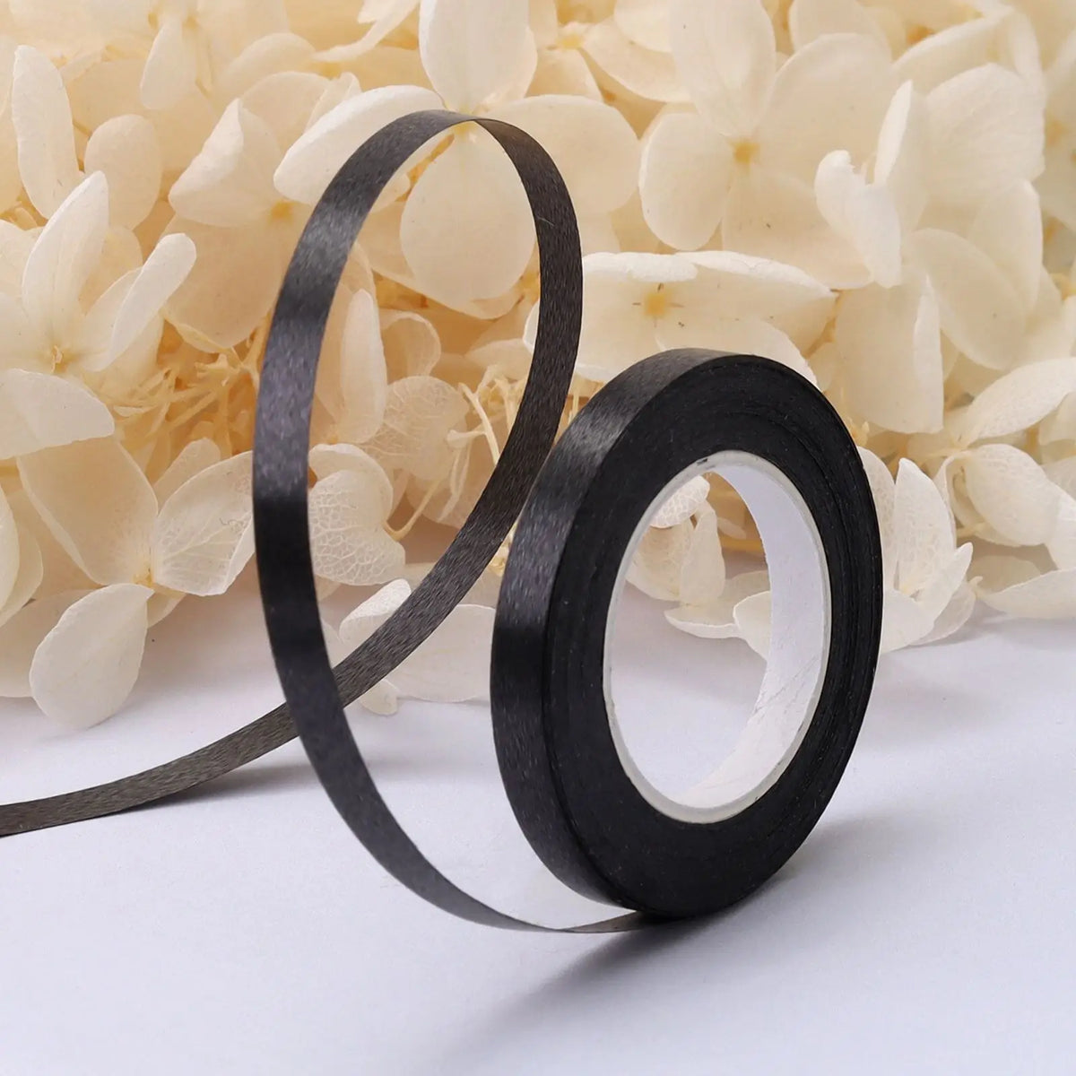 A roll of black satin balloon ribbon is shown against a background of light yellow flowers.