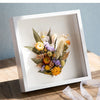 A white wooden shadow box frame containing dried flowers and greenery.