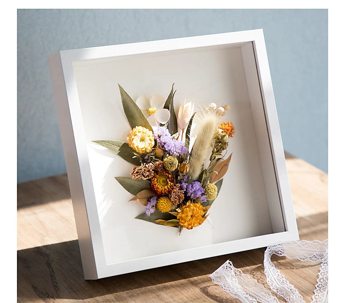 A white wooden shadow box frame containing dried flowers and greenery.