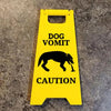 A yellow yard sign with black text warning of dog vomit and caution, featuring a silhouette of a dog.