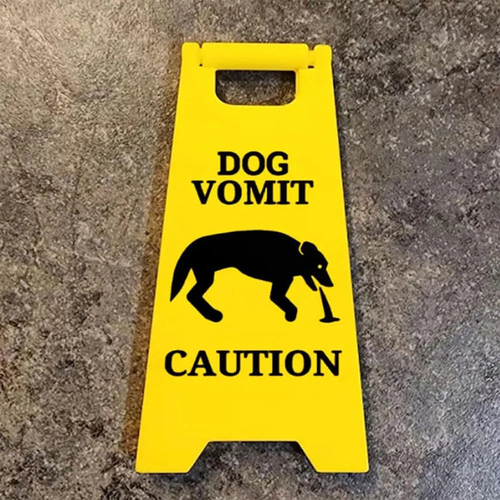 A yellow yard sign with black text warning of dog vomit and caution, featuring a silhouette of a dog.