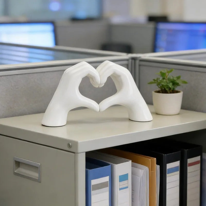 A white heart shape sculpture made of two hands rests on a light gray shelf next to some office supplies.