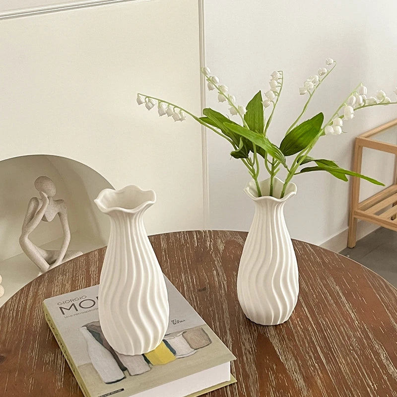 A white ceramic bud vase with vertical ridges sits on a wooden table next to a book.