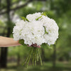 A hand holds a bouquet of large white artificial hydrangea flowers with green stems against a blurred green outdoor backgroun