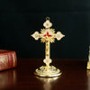 A golden metal Jesus cross ornament with a decorative base, displayed on a dark wooden surface.