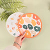 A hand holds an orange portable foldable fabric hand fan with a floral pattern against a light green background.