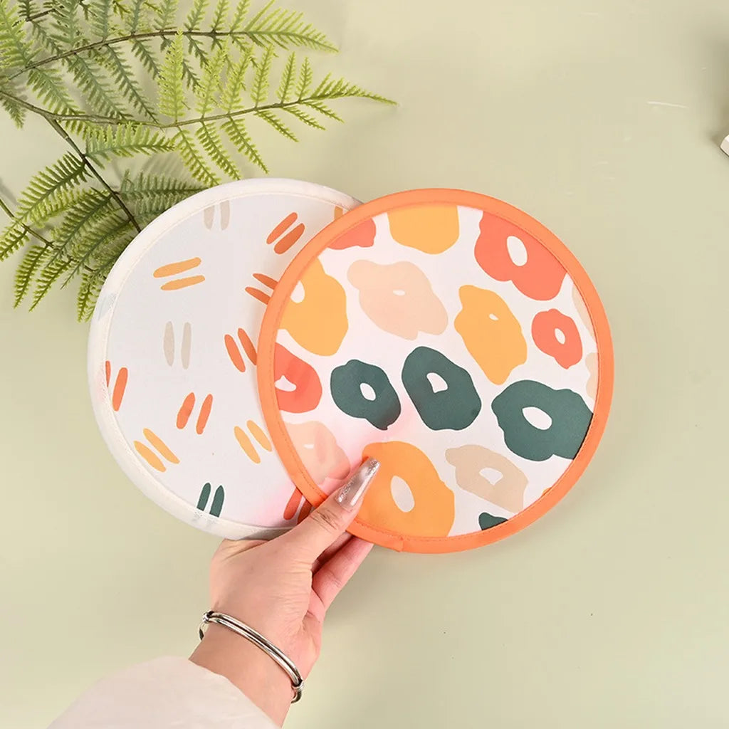 A hand holds an orange portable foldable fabric hand fan with a floral pattern against a light green background.