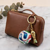 A brown leather pouch with a gold zipper and a FIFA trophy keychain resting on a white marble surface.