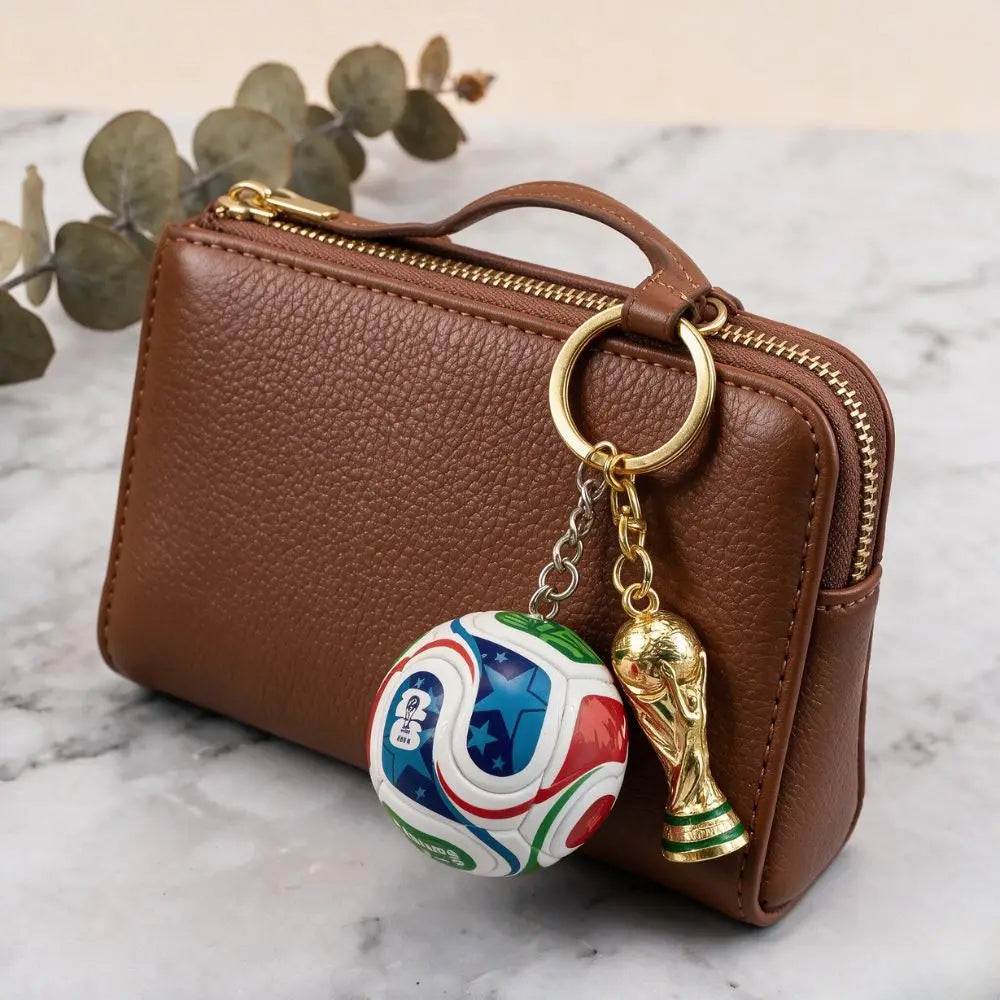 A brown leather pouch with a gold zipper and a FIFA trophy keychain resting on a white marble surface.
