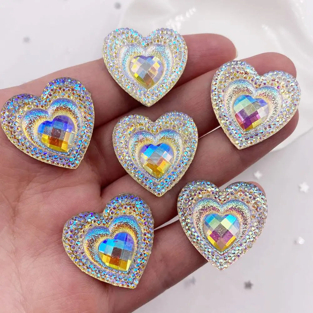 A hand holds a set of twelve heart-shaped AB black peacock resin rhinestone flatbacks.