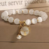 A bracelet made of small, round white natural stones with a gold-toned charm and pearl drop on a wooden surface.