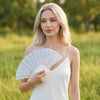 A blonde woman models a white dress and holds a glitter folding fan with transparent ribs outdoors.