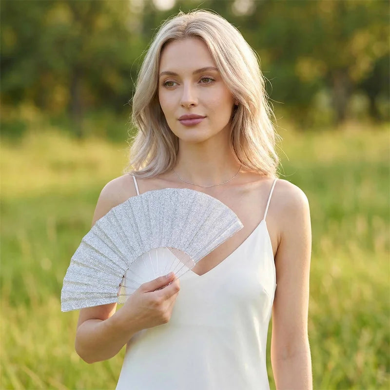 A blonde woman models a white dress and holds a glitter folding fan with transparent ribs outdoors.
