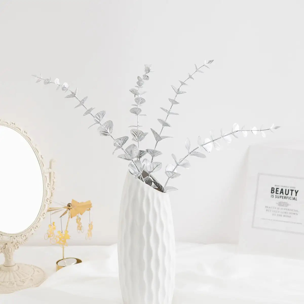 A vase filled with artificial eucalyptus leaves on a white surface next to a mirror and a framed text.