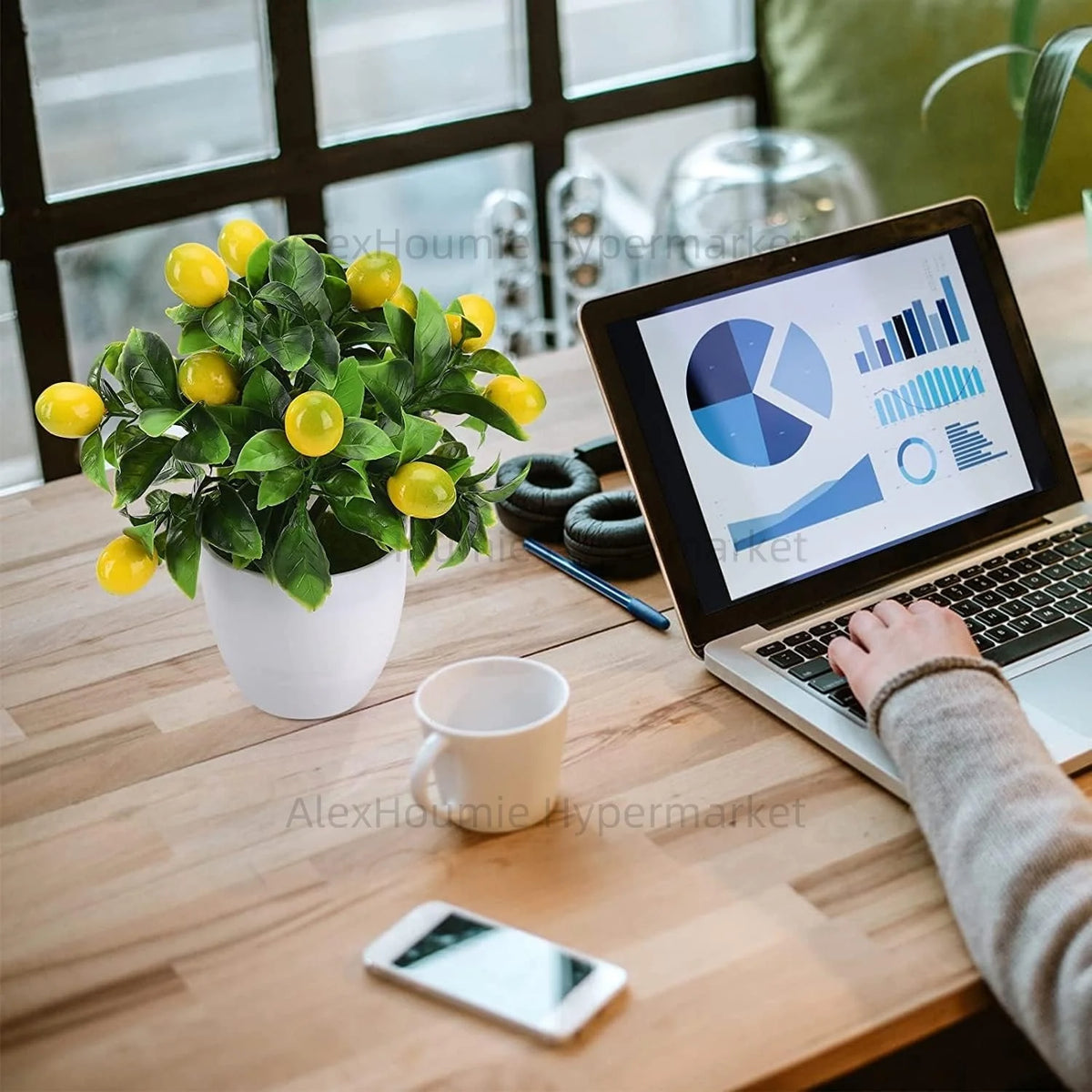 A laptop displaying a chart is open on a wooden table next to an artificial lemon tree bonsai, a white mug, and a smartphone.