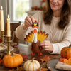 A woman holds a felt turkey costume with a goose statue on a wooden table surrounded by autumn leaves and pumpkins.