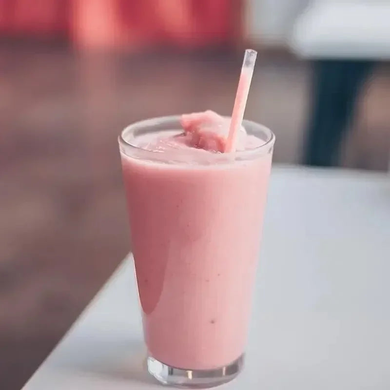 A glass of pink smoothie with a plastic straw is shown on a white surface.
