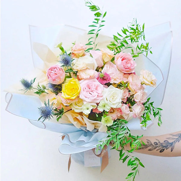 Un ramo de rosas rosa y crema con follaje envuelto en papel floral con borde dorado.