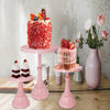 A vibrant red frosted cake is displayed on a pink metal stand next to other desserts and flowers.