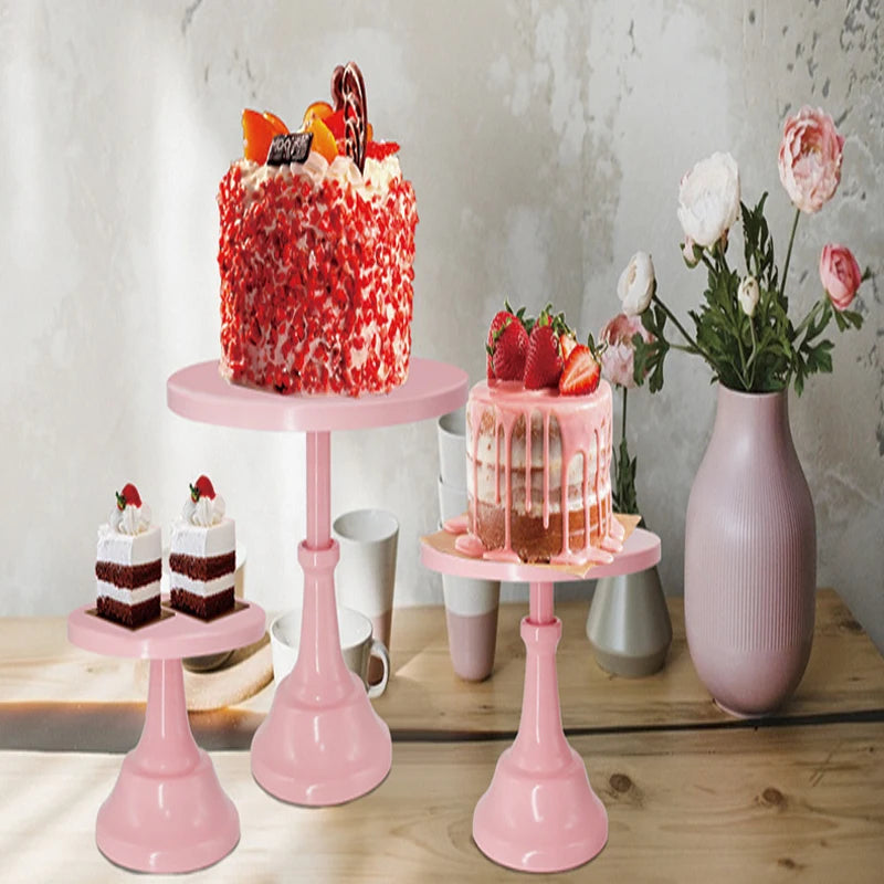 A vibrant red frosted cake is displayed on a pink metal stand next to other desserts and flowers.
