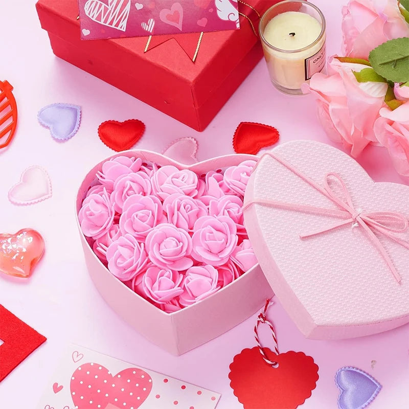 Una caja con forma de corazón rosa llena de rosas artificiales rosas, junto a una caja de regalo roja y flores rosas.