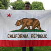 A person holds up a large white flag featuring a brown bear silhouette on green land and the text CALIFORNIA REPUBLIC.