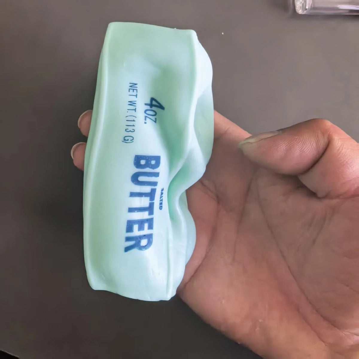 A light green squeeze butter product tube is held in a hand against a gray background.