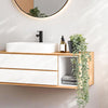 Un lavabo de salle de bain avec évier blanc, robinet noir et meuble en bois avec plantes d'eucalyptus artificielles.