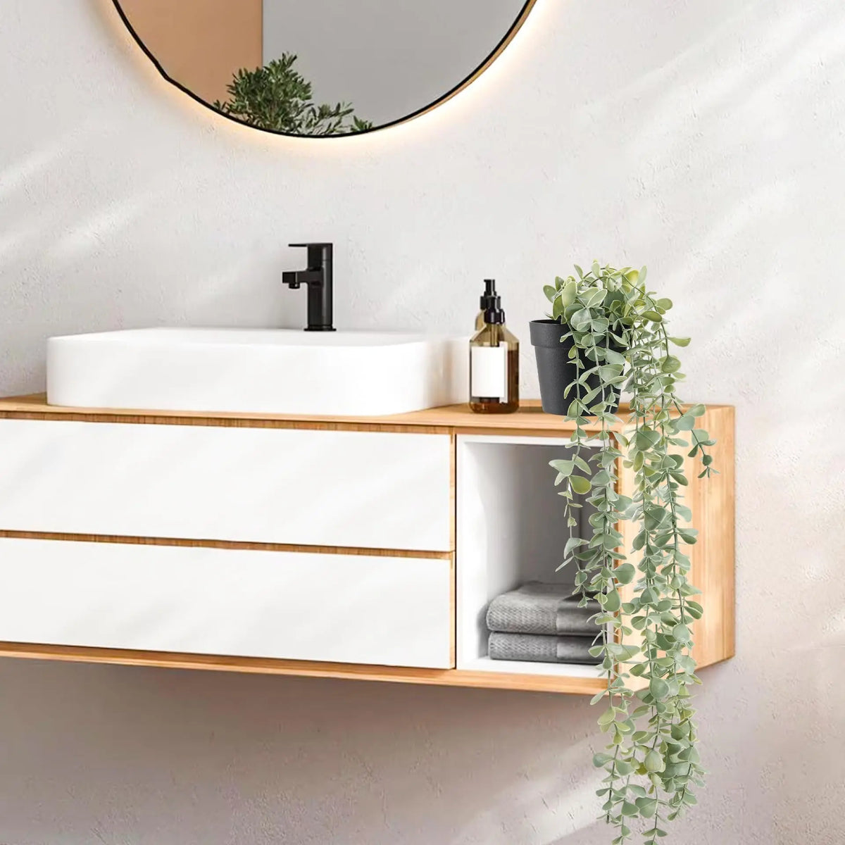 Un lavabo de salle de bain avec évier blanc, robinet noir et meuble en bois avec plantes d'eucalyptus artificielles.