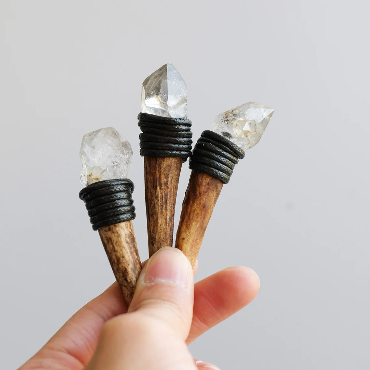 A hand holds three natural crystalHerkimer scepter wands with dark wrappings and clear crystals.