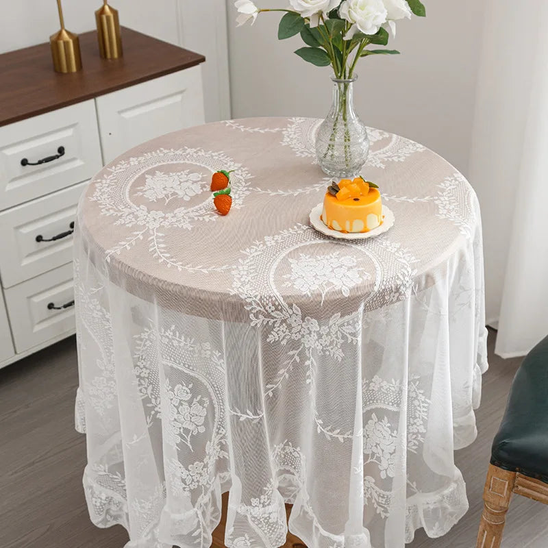 Elegant white floral lace tablecloth covering a round table set with a vase of white flowers.