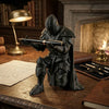 A dark, 3D printed medieval knight figurine holding a pen on a wooden desk next to books and an oil lamp.