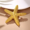 A gold glitter starfish brooch pin set lying on a beige surface with soft shadows.
