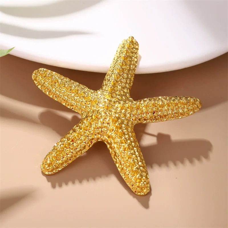 A gold glitter starfish brooch pin set lying on a beige surface with soft shadows.