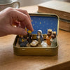 A hand places a mini nativity set featuring figures like Mary, Joseph, and animals into a gold tin storage box.