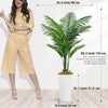 A tall artificial palm tree in a white pot displayed next to a woman wearing beige overalls.