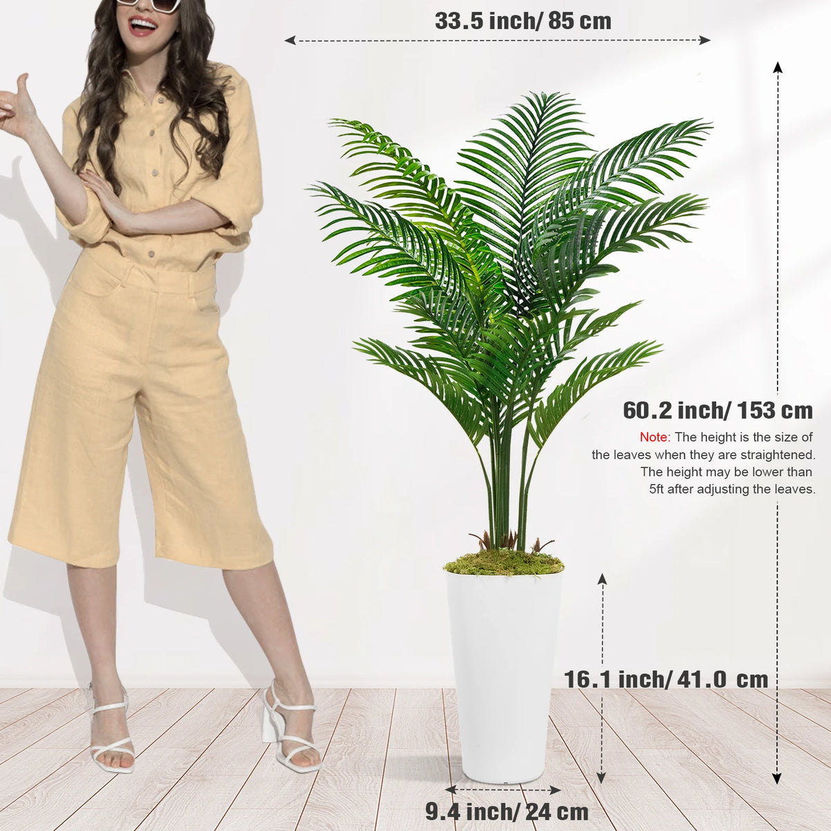 A tall artificial palm tree in a white pot displayed next to a woman wearing beige overalls.