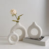 A white ceramic circular vase holding a single pale pink rose, displayed on a light surface.