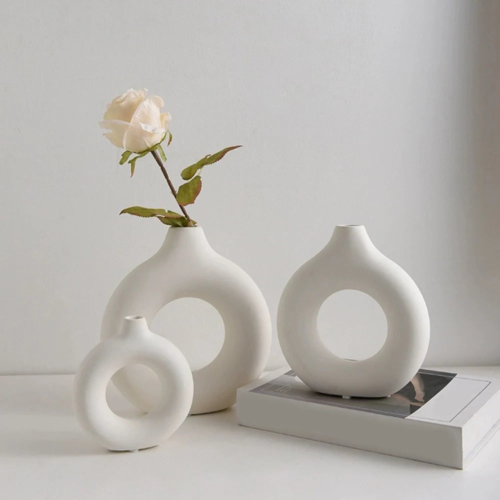A white ceramic circular vase holding a single pale pink rose, displayed on a light surface.
