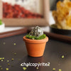 A small, moss-covered piece of chalcopyrite crystal is planted in an orange terracotta pot on a dark surface.