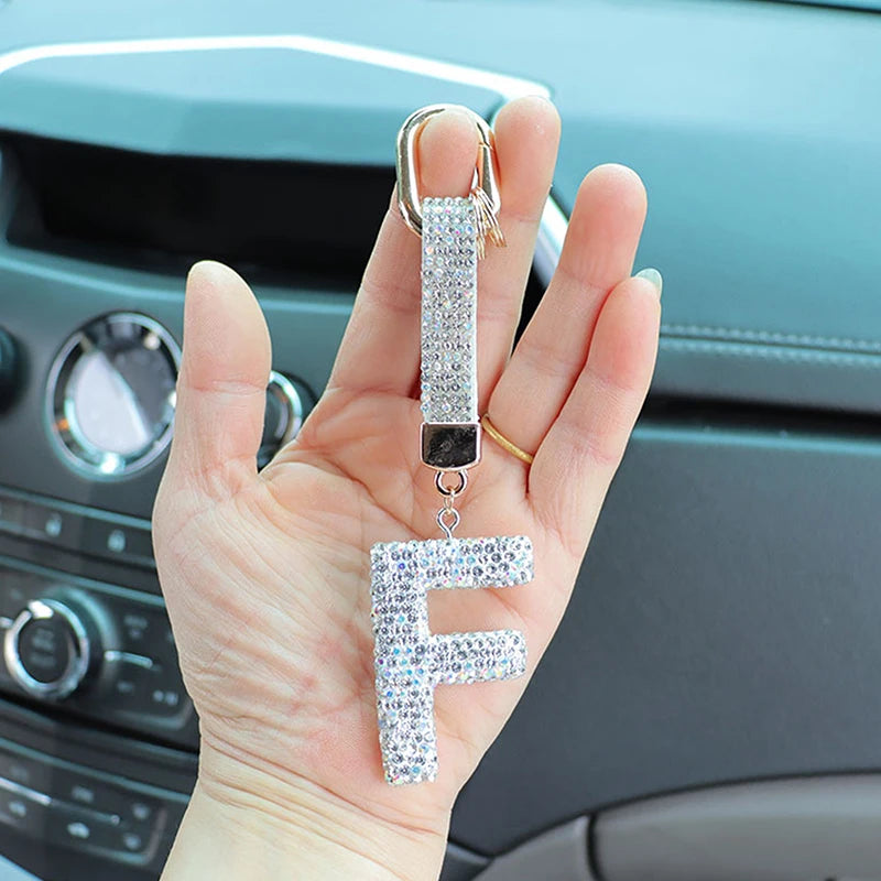 A hand holds a crystal letter keychain charm shaped like the letter F against a dark car interior.