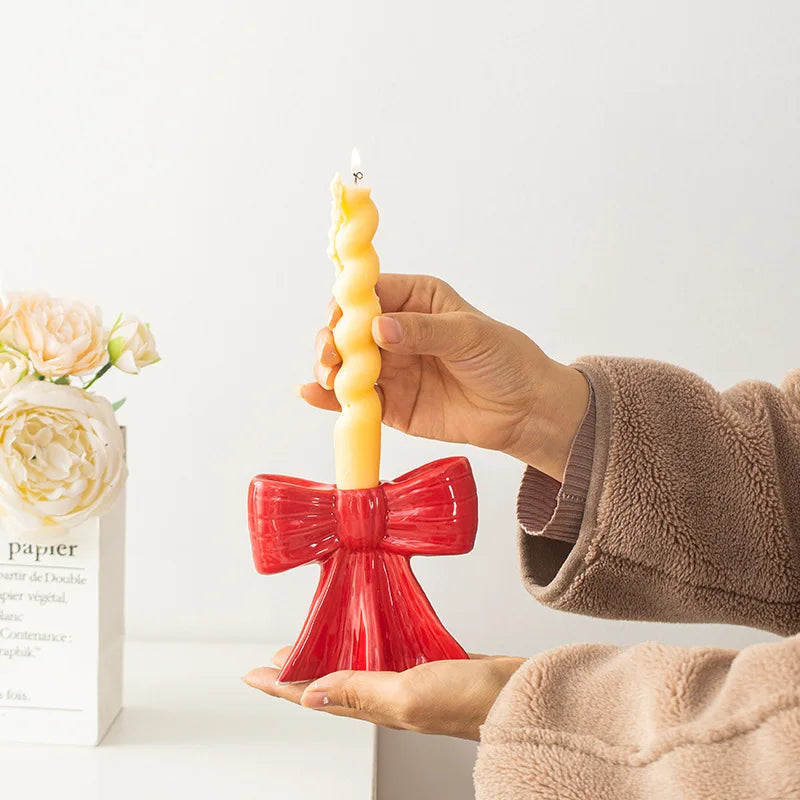 A hand holds a yellow candle in a red ceramic bow holder against a white background.