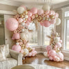 A pink and gold balloon garland arch decoration featuring various sized balloons and dried floral accents in a bright room.