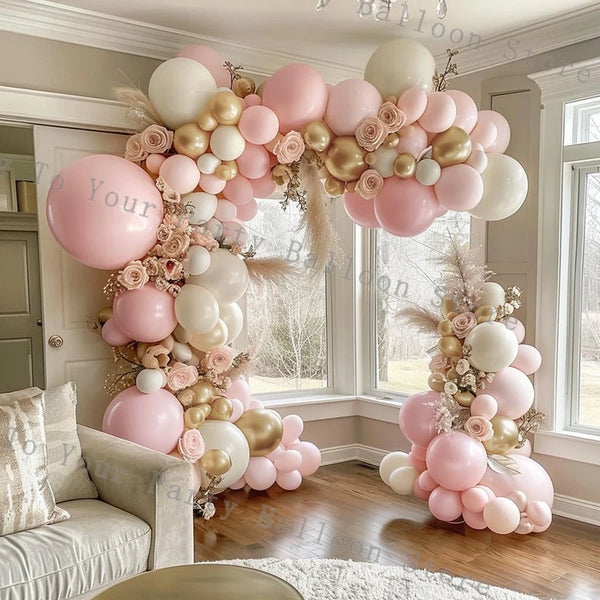 A pink and gold balloon garland arch decoration featuring various sized balloons and dried floral accents in a bright room.