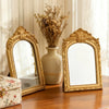 A pair of ornate gold-framed vanity mirrors sit on a wooden table next to a vase of dried flowers.