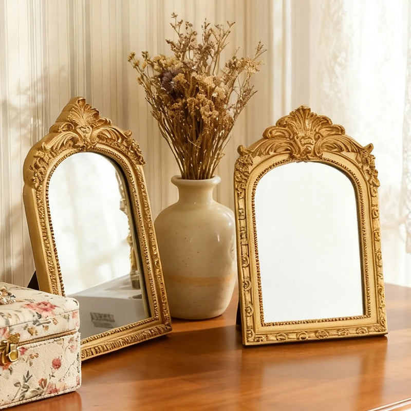 A pair of ornate gold-framed vanity mirrors sit on a wooden table next to a vase of dried flowers.