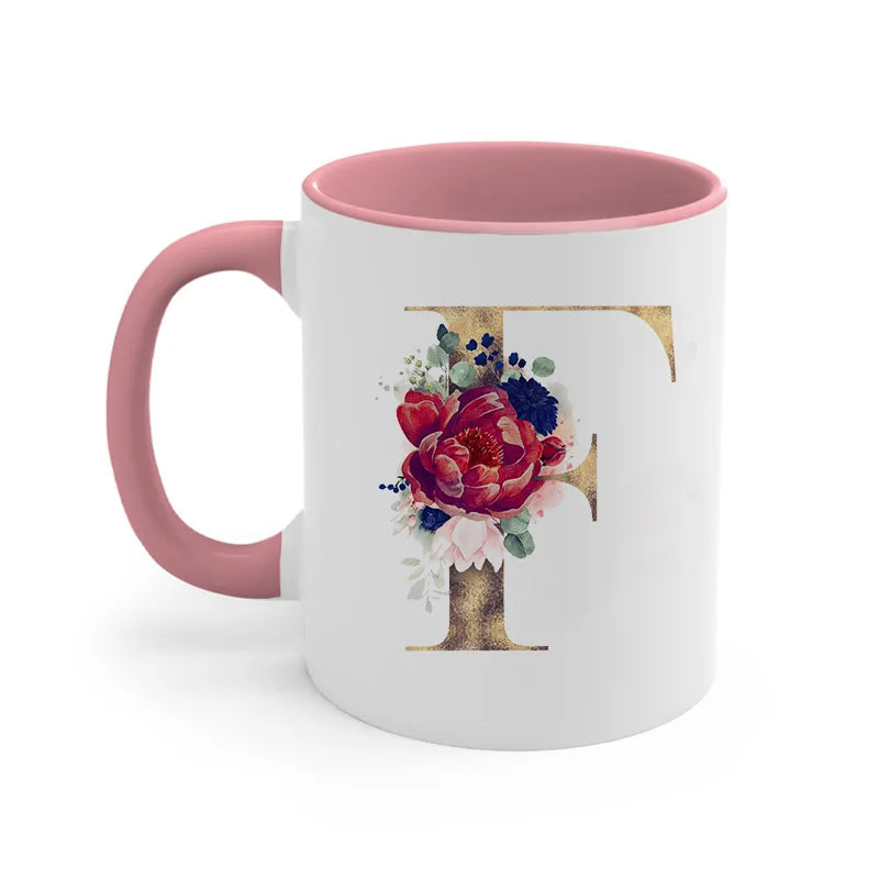 A pink ceramic coffee mug featuring a gold letter initial decorated with colorful flowers and greenery.