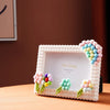 A white photo frame decorated with colorful foam blocks and greenery, featuring a pink floral arrangement.